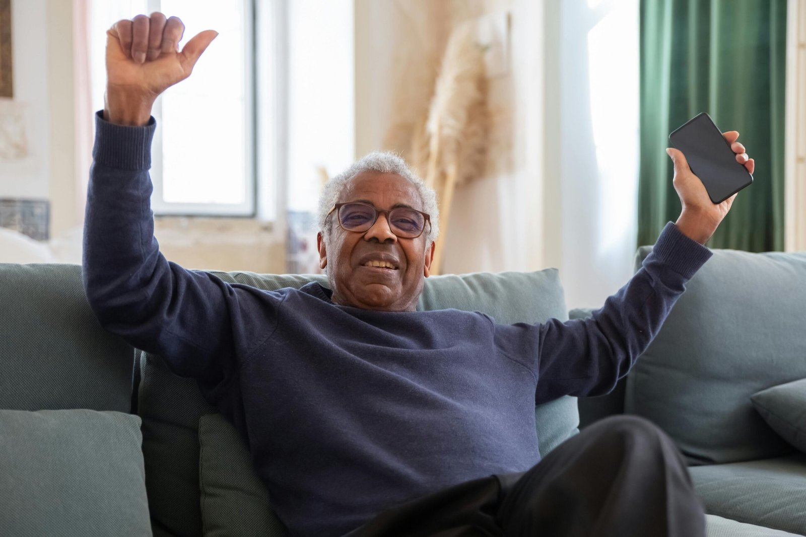 Senior man joyfully sitting on a couch holding a smartphone indoors.