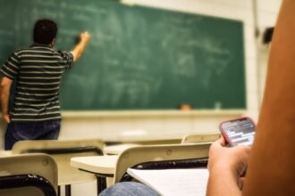 Student texting in a classroom while teacher is writing on the blackboard.