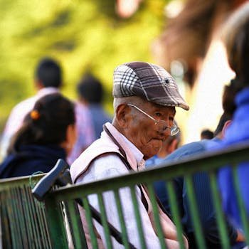 An elderly man in a hat sits pensively on a park bench, surrounded by blurred figures.