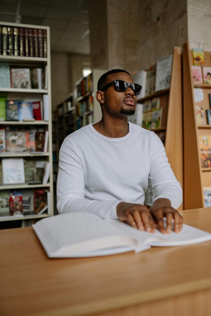 A blind man reads a braille book at a library desk, engaging in tactile reading.